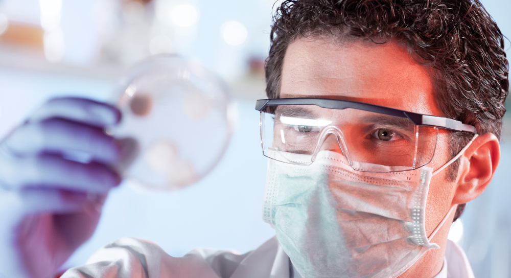 man in safety goggles and mask holding up and inspecting a petri dish